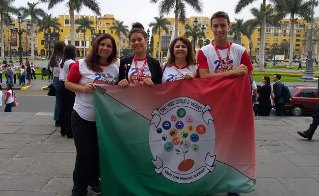 Três alunos e uma professora em pé, atrás de uma bandeira
