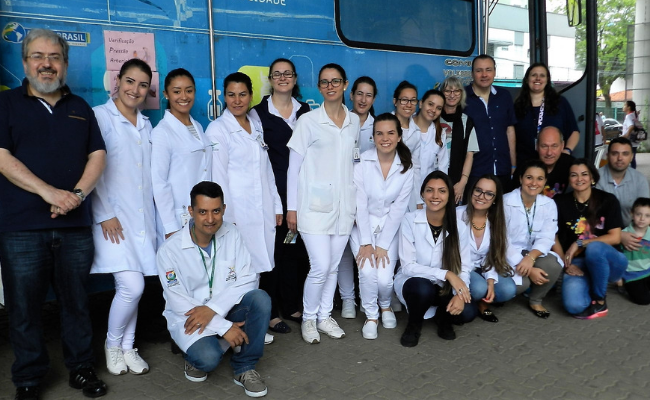Com o ônibus atrás, equipe de profissionais da saúde lado a lado, sendo um grupo em pé e outro agachado