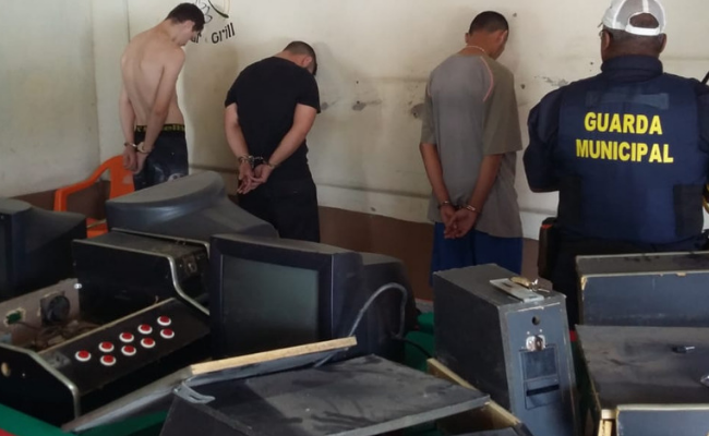 Quatro homens escorados na parede com os braços para cima e um guarda municipal de costas. Máquinas caça-níqueis em cima de uma mesa