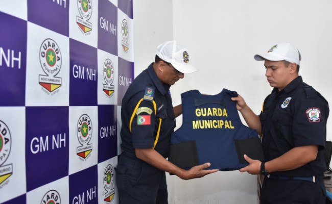 Dois guardas municipais fardados segurando um colete azul escrito Guarda Municipal em amarelo
