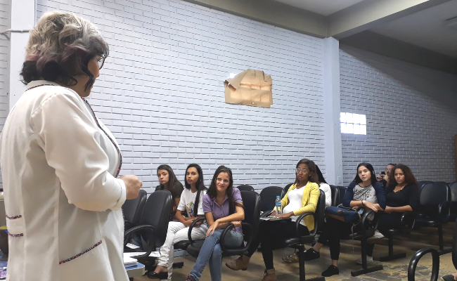 Professora em pé e alunas sentadas a sua frente