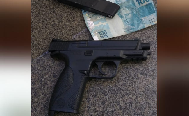 Em cima de uma mesa, um simulacro preto de uma pistola e notas de dinheiro