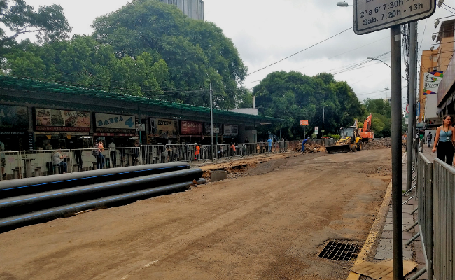 Uma rua com uma máquina trabalhando e canos pretos depositados no lado esquerdo. À esquerda também, as oito bancas que compõem o local. 