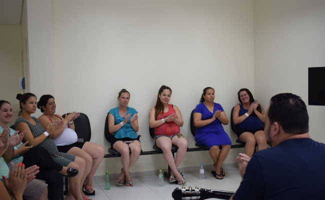 Um homem de costas tocando violão e um grupo de mulheres sentandas no entorno