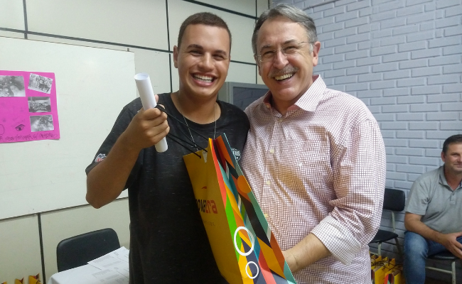 Dois homens em pé, abraçados, sendo um aluno vestindo preto à direita e o secretário Roberto à direita