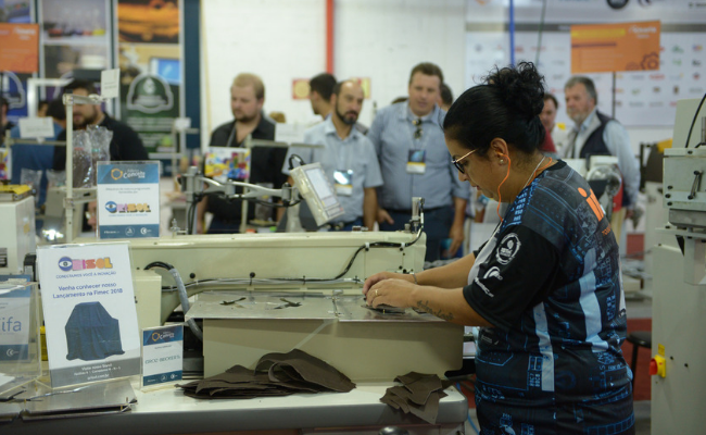 Uma mulher em pé manuseia o solado de uma sapato em um máquina. Ao fundo, pessoas assistem o processo