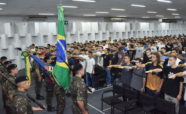 Em um auditório, jovens em pé com o braço direito esticado em direção a uma bandeira do Brasil