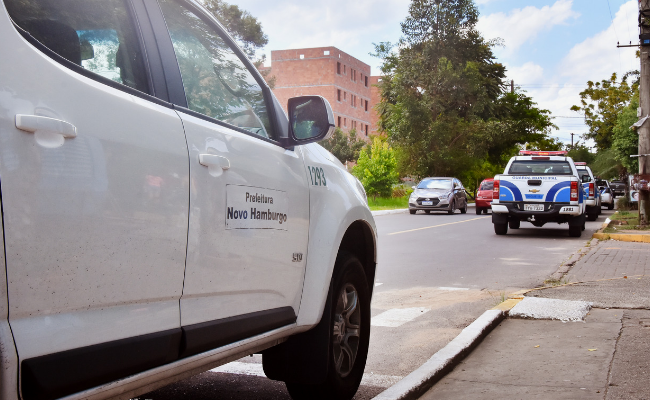 Carro branco da Prefeitura em uma rua com uma viatura da Guarda Municipal ao fundo
