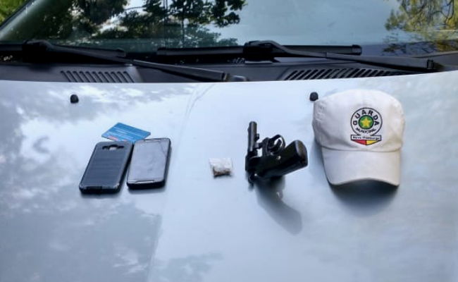 Em um capô de um carro, dois telefone, um pequeno pacote branco com droga, um revólver, e um boné da Guarda Municipal