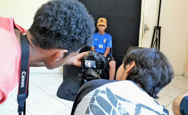 Ao fundo, um jovem sentado com um pano preto atrás. A frente, dois jovens olham o visor de uma câmera em um tripé