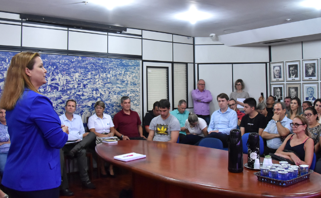 Em uma sala, prefeita em pé, uma mesa no centro e comerciantes sentados