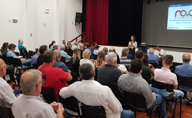 Ao fundo, a secretária em pé. Público sentado em um auditório