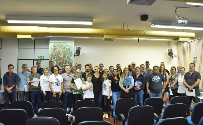 Em um auditório, grupo de pessoas em pé ao fundo com a prefeita no meio