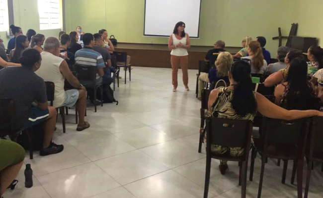 Ao fundo, em pé, a secretária Maristela. Coordenadores sentados em cadeiras distribuídas de um lado e outro de uma sala, com um corredor no meio