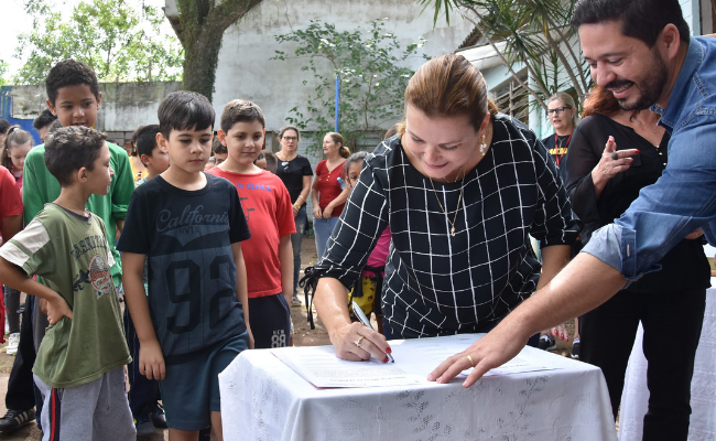Prefeita assinando um papel em cima de uma mesa com uma toalha branca