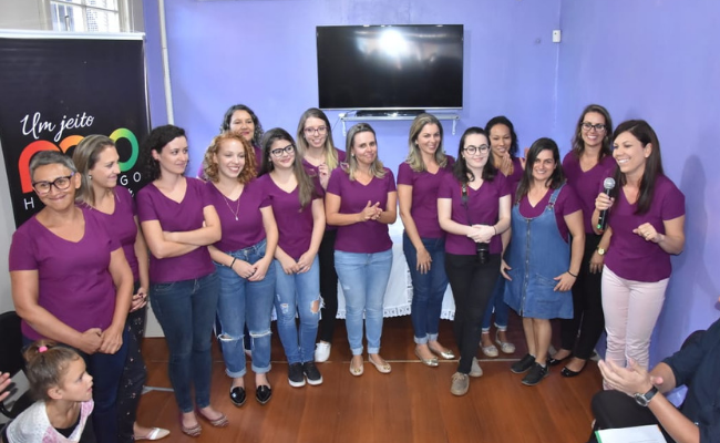 Em pé, a coordenadora Elis Regina ao microfone, com a equipe do CREAS Viva Mulher II vestindo um camiseta rosa ao seu lado