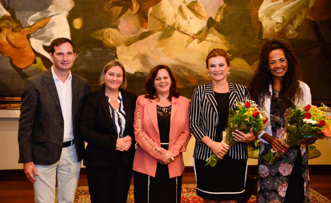 Um homem e quatro mulheres em pé. As duas últimas mulheres da esquerda para a direita seguram um buquet de flores