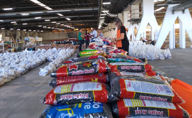 Em uma mesa, sacos de feijão empilhados e ao fundo sacolas brancas com alimentos no chão