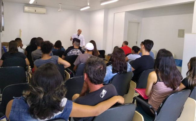 Em uma grande sala, um médico em pé fala para uma platéia sentada
