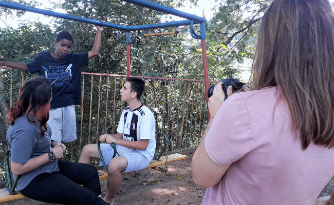 Uma menina com uma câmera na mão, filma três jovens conversando, dois deles sentados, sendo uma mulher e um homem, e um outro homem em pé