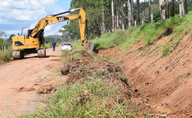 Uma máquina amarela abre um valo em uma estrada de terra. Ao fundo, o secretário Raizer em pé no meio da estrada, atrás da máquina