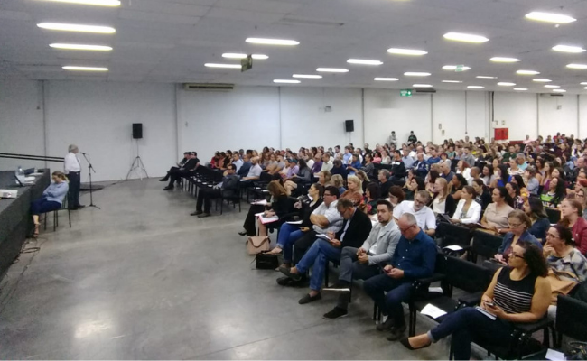 Um homem no microfone ao fundo e platéia sentada a sua frente