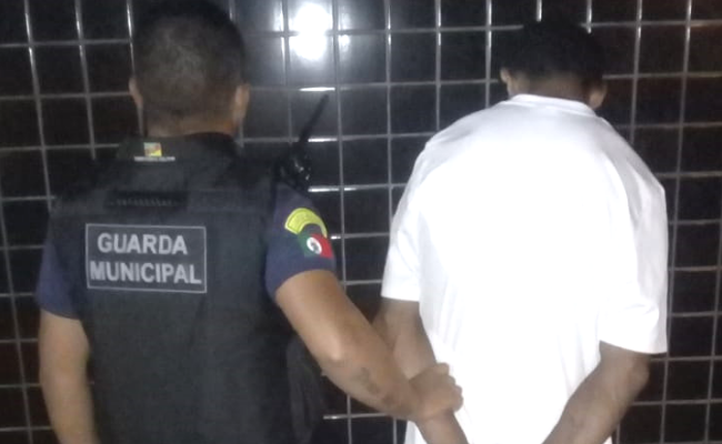 Do lado esquerdo, de costas, um Guarda Municipal fardado. Do outro lado, também de costas, o preso vestindo branco
