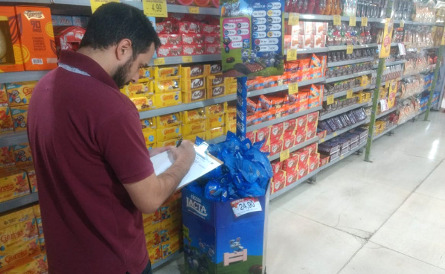 Em um corredor de supermercado, homem com uma planilha na mão olha chocolates em uma prateleira