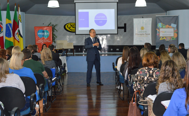 Ao fundo, um homem em pé palestra. Público sentado a sua frente, dividido em dois blocos, um a esquerda e o outro à direita, com um corredor no meio