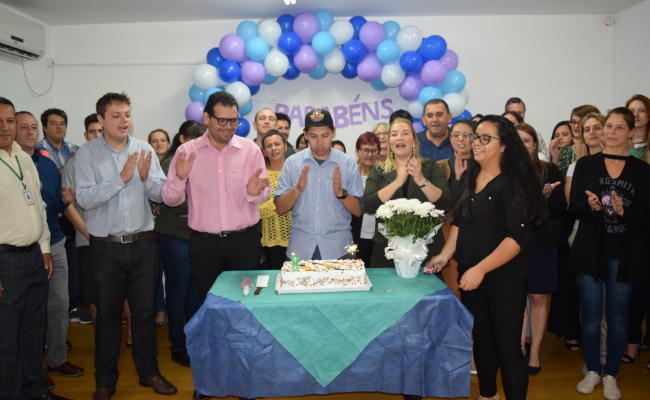 Com uma mesa à frente e um bolo em cima, grupo de pessoas atrás da mesa cantando parabéns