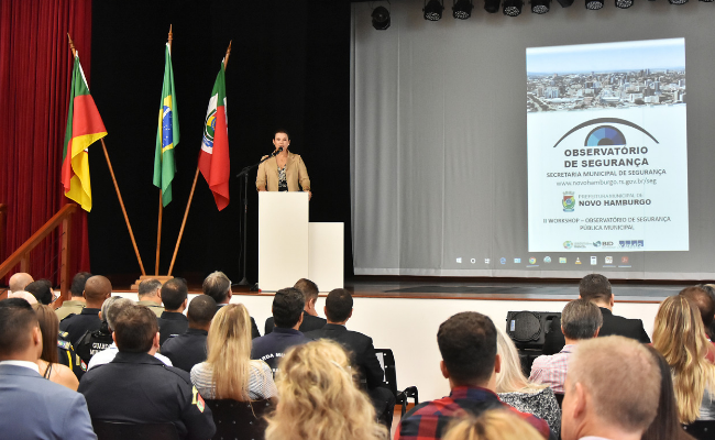 Em um palco, a prefeita fala em um pulpito e o público sentado
