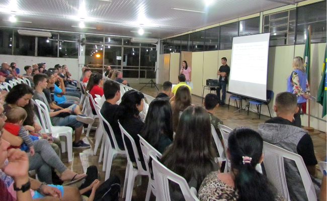 Jovens sentados em cadeiras enquanto uma professora fala ao microfone mostrando uma projeção na parede