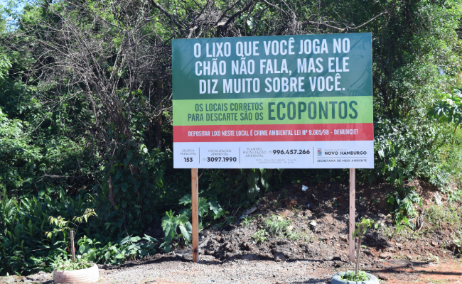 Em uma área de terra, uma placa com a frase “O lixo que você joga no chão não fala, mas ele diz muito sobre você. Os locais corretos para o descarte são os ECOPONTOS”