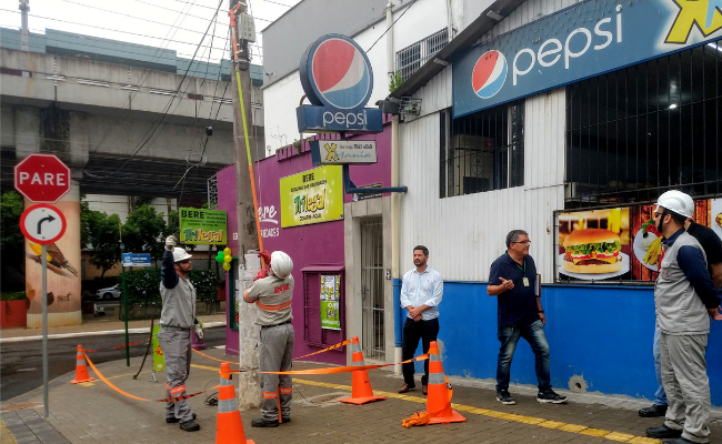 Em um poste, dois homens com uniforme da RGE mexem em um poste. Ao lado, o secretário Raizer acompanha o serviço 