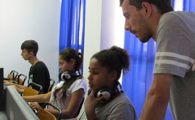 Três alunos sentados à frente de computadores e um professor auxilia este estudantes em pé ao lado