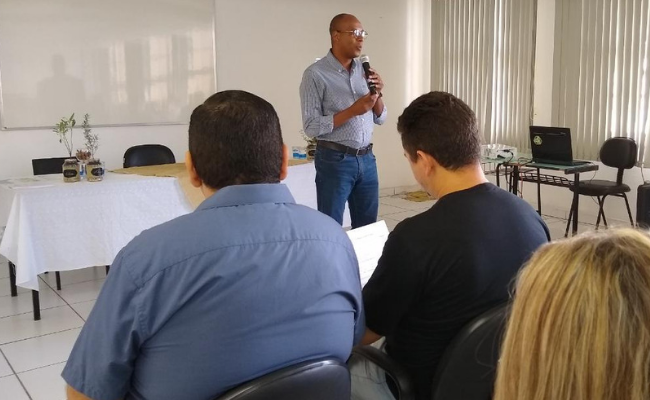 Em uma sala, secretário Daniel em pé ao microfone fala para uma platéia sentada