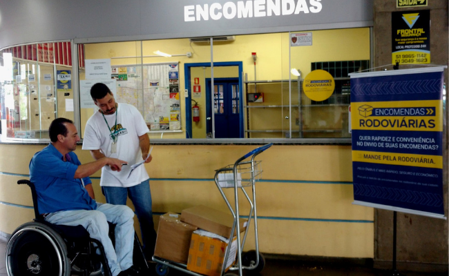 Em frente ao balcão de encomendas, um homem em uma cadeira de rodas e outro homem em pé ao seu lado conferem em uma planilha as encomendas, que estão em caixas ao seu lado 