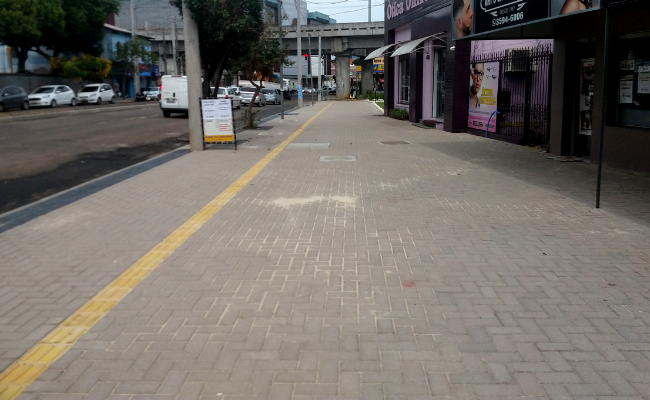 Calçada de um rua com blocos. Ao lado direito, lojas, e ao lado esquerdo, a rua