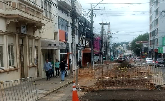 À esquerda, lojas com pessoas caminhando na calçada. À direita, a rua em obras fechada com gradis
