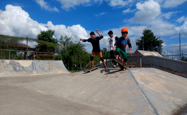 Em um pista de skate, três jovens em cima do skate descem de um obstáculo