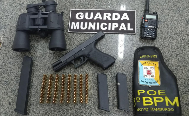 Em cima de uma mesa, uma arma, cartuchos de bala, um rádio comunicador, um binóculos e as insigna da Guarda Municipal e da Brigada Militar