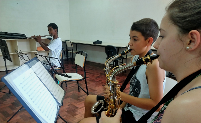 Três alunos sentados lado a lado tocando saxofone