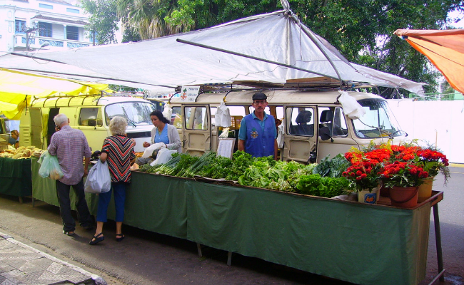 Frutas e verduras expostas em uma mesa, com um vendedor e duas pessoas comprando. Uma Kombi está estacionada atrás