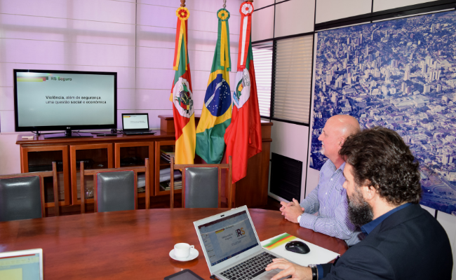 Em uma mesa, um homem sentado na ponta e outro ao lado com um computador a sua frente. À esquerda, uma televisor em cima de um balcão