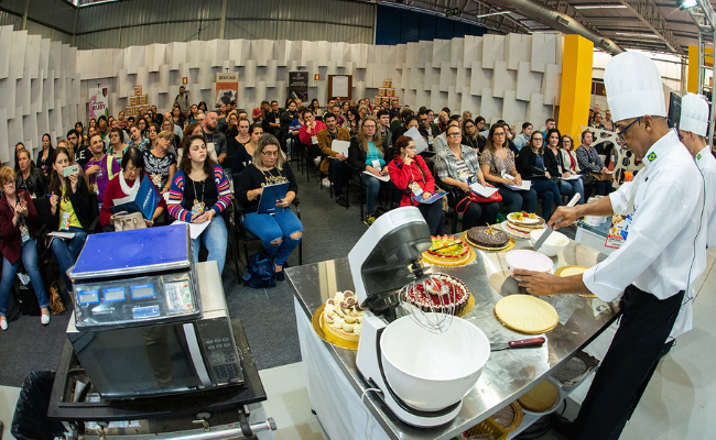 Em um auditório, público sentado. Em uma palco, um chefe de cozinha cozinha