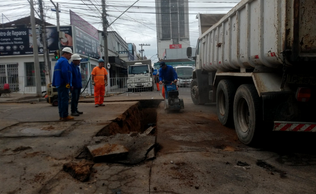 Em uma rua, três homens trabalham em um buraco retângular. Ao lado, um caminhão estacionado