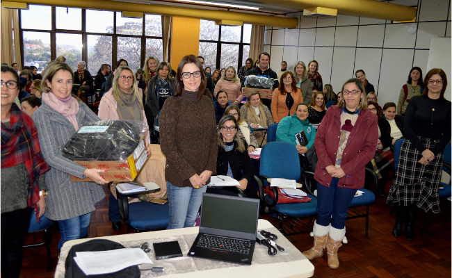 Em uma sala, uma mesa com um computador, e atrás grupo de mulheres, umas em pé e ouras sentadas