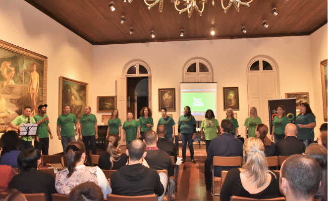 Coral em pé ao fundo e grupo de pessoas sentadas a sua frente