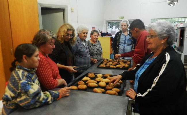 Pessoas no entorno de uma mesa. Na mesa, formas com pães