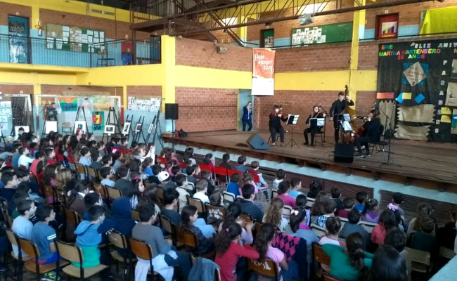 Em um palco, três pessoas tocam instrumentos. Em sua frente, uma platéia sentada